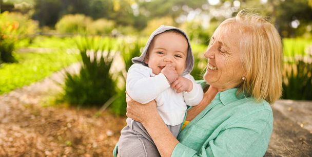 Beautiful happy smiling senior elderly woman holding on hands cute little baby boy. Grandmother and grandson having fun time together at tropical summer day in park. Multigenerational family concept - Photo, Image