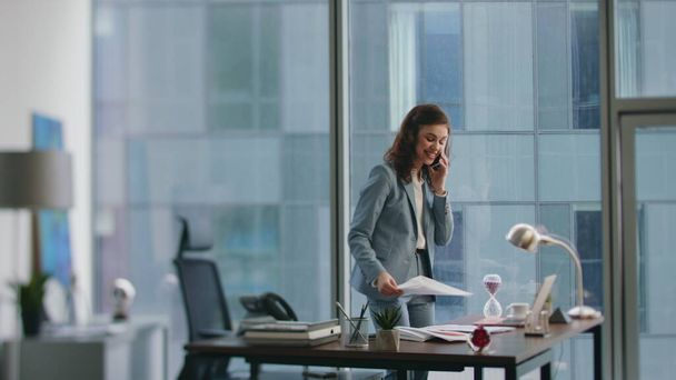 Жінка-менеджер розмовляє смартфоном, що стоїть біля робочого столу. Щаслива усміхнена бізнес-леді, яка телефонує партнерам, ділиться успіхом роботи. Весела задоволена дівчина перестає розмовляти телефоном, шукаючи документ на офісному столі - Фото, зображення