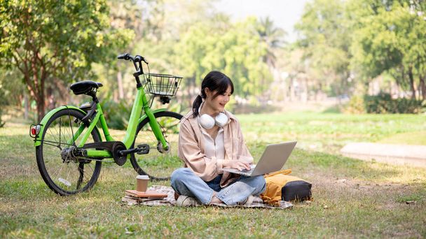 Щаслива і чарівна молода азіатка працює дистанційно в зеленому парку, використовуючи свій ноутбук. Студентка коледжу робить домашнє завдання онлайн, відпочиваючи в кампусному парку. - Фото, зображення