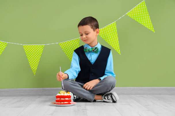 Petit garçon mignon avec gâteau d'anniversaire et guirlande assis près du mur vert - Photo, image