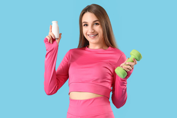 Sporty νεαρή γυναίκα με μπουκάλι νόστιμο γιαούρτι και dumbbell σε μπλε φόντο - Φωτογραφία, εικόνα