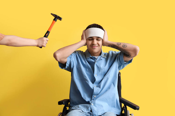 Joven con conmoción cerebral en silla de ruedas y martillo sobre fondo amarillo - Foto, Imagen