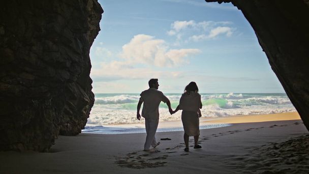 岩の影から海岸を歩くカップルを見る. 若い愛する配偶者は,幸せな気分で抱擁する黄金の砂の海岸を踏みます. 海の波の夏休みに行く手を握っているリラックスした既婚女性 - 写真・画像