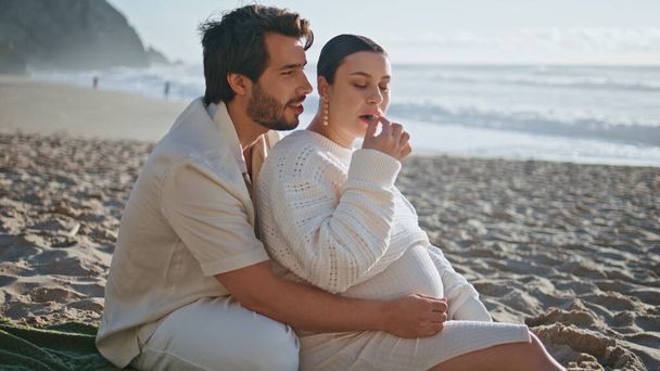妊娠中の家族がピクニックに座って,夕方の海岸で新鮮な果実を食べる. 美しい海の自然を楽しむ大きな妻の腹を抱擁する愛する夫. 赤ちゃんを待っているロマンチックなカップルリラックス週末. - 写真・画像