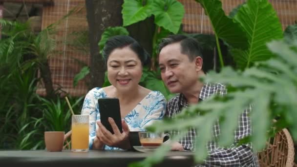 Střední záběr veselých dospělých asijských manželek při pohledu na fotografie jejich vnoučat na smartphonu sedí u stolu ve venkovní kavárně - Záběry, video