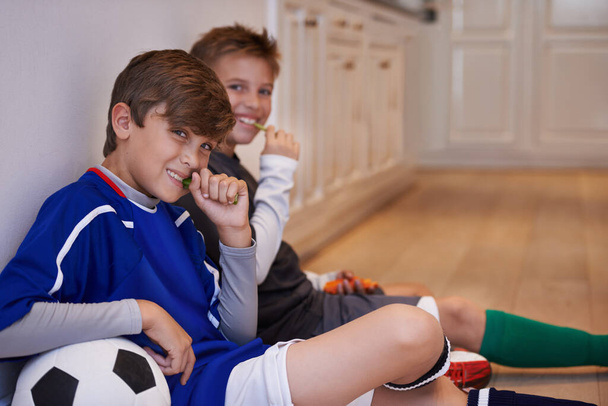 Children, portrait and soccer ball for sport relax while eating food in apartment or hungry, practice or friends. Male people, face and football training in kitchen for nutrition snack, break or kit. - Photo, Image