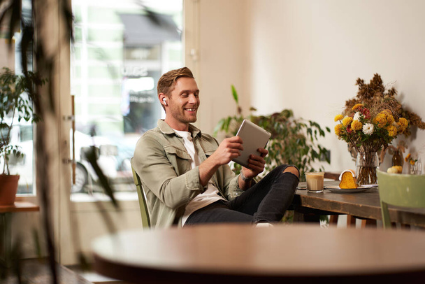 Portret szczęśliwego, zrelaksowanego młodzieńca siedzącego samotnie w kawiarni, noszącego słuchawki bezprzewodowe, grającego w gry wideo na tablecie, śmiejącego się i uśmiechającego, patrzącego na swoje urządzenie. Pojęcie stylu życia i ludzi. - Zdjęcie, obraz