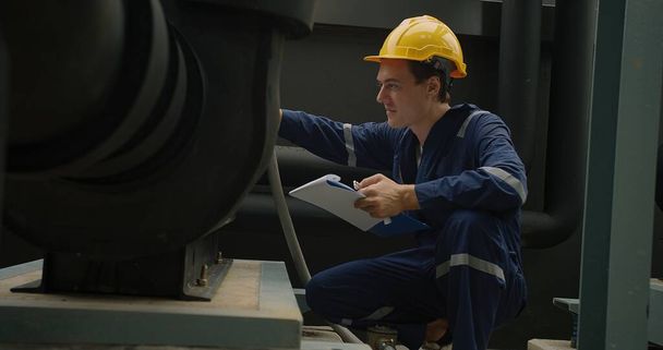 Teollisuustyöntekijä sinisissä haalareissa ja keltainen kypärä tarkastelevat huolellisesti koneita leikepöydällä. - Valokuva, kuva
