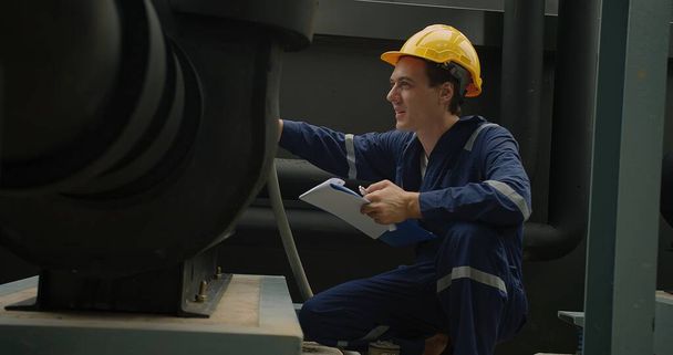 Teollisuustyöntekijä sinisissä haalareissa ja keltainen kypärä tarkastelevat huolellisesti koneita leikepöydällä. - Valokuva, kuva