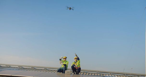 Güvenlik yelekleri ve başlıkları olan iki mühendis, açık mavi gökyüzüne doğru uçan bir İHA ile güneş panellerini inceliyor. - Fotoğraf, Görsel