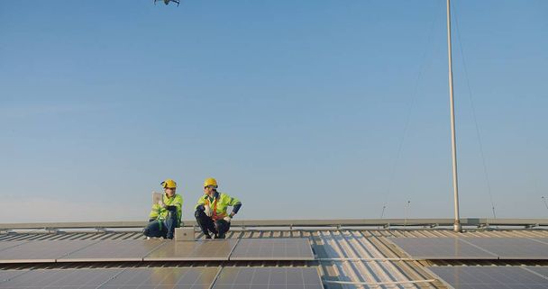 Dos ingenieros con chalecos de seguridad y sombreros duros inspeccionan paneles solares con un dron volando sobre un cielo azul claro - Foto, imagen
