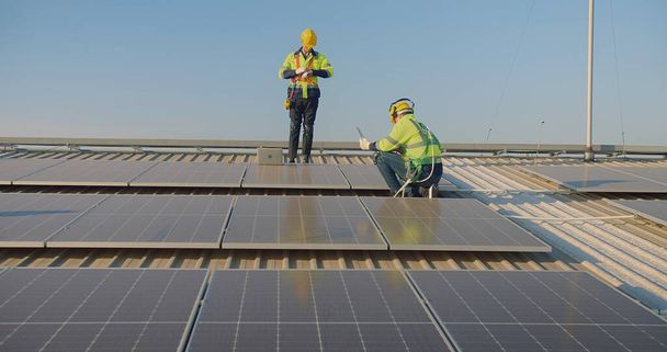 Güneş paneli teknisyenleri yansıtıcı teçhizatlı işçiler arka planda gün batımında geniş bir güneş dizisi üzerinde bir dizüstü bilgisayar üzerinde tartışıyorlar. - Fotoğraf, Görsel