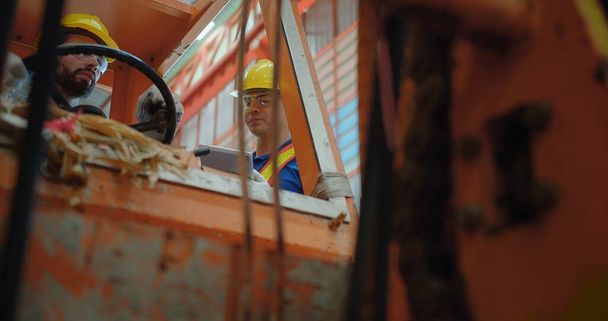 Οι εργαζόμενοι στις κατασκευές με κίτρινα σκληρά καπέλα και γιλέκα ασφαλείας χειρίζονται μηχανήματα ανελκυστήρων μέσα σε ένα βιομηχανικό περιβάλλον, εστιάζοντας στην εργασιακή ασφάλεια και την ομαδική εργασία - Φωτογραφία, εικόνα