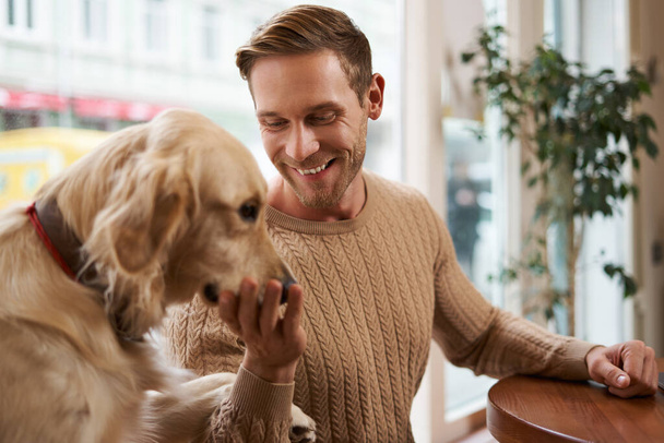 Portrait of handsome smiling blond guy sits in cafe and pets his dog, touches golden retriever. - Photo, Image
