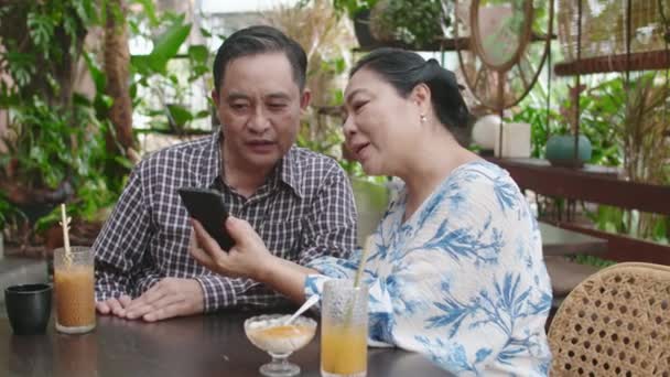 Střední záběr starší asijské ženy vyučování manžel používat mobilní telefon sedí u stolu s nápoji v kavárně - Záběry, video