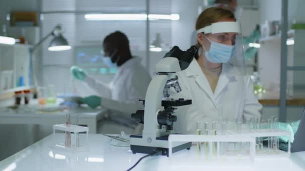 Taille omhoog van vrouwelijke wetenschapper die gezichtsschild, masker, handschoenen en labjas gebruikend microscoop terwijl het onderzoek van vloeistof van reageerbuis, het werken met diverse mannelijke collega 's in laboratorium - Video