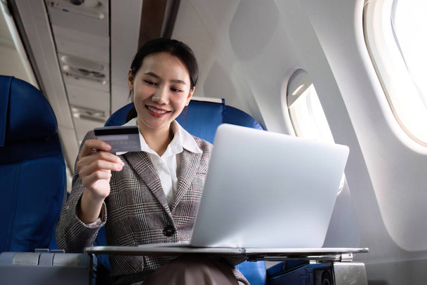 Mujer asiática joven sosteniendo compras con tarjeta de crédito en línea utilizando el sitio web en el ordenador portátil Sentado cerca de la ventana en primera clase en el avión durante el vuelo, viaje y concepto de negocios.. - Foto, imagen