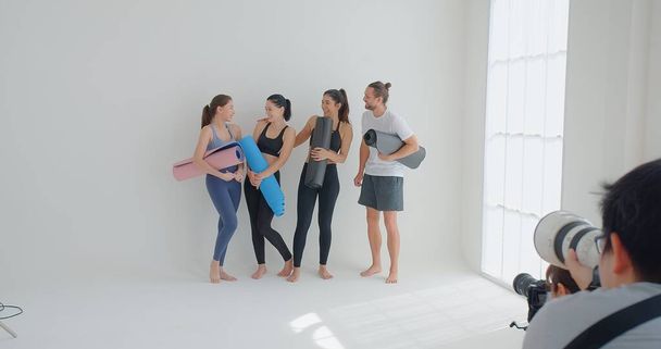 Fotograf fotografiert Gruppe von Yoga-Studenten, die beim Yoga-Kurs an die Wand gelehnt stehen, Menschen in Sportbekleidung halten Yogamatten in der Hand, die glücklich und gesund aussehen, Wohlbefinden, Sport-Wellness-Konzept - Foto, Bild