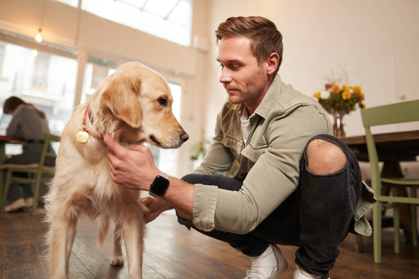 Retrato de homem feliz passar o tempo com seu cão no café pet-friendly, jogando e acariciando golden retriever, se divertindo, dando-lhe um deleite. - Foto, Imagem