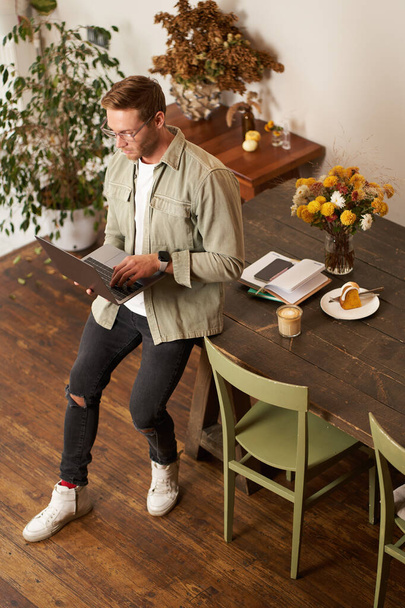 Porträt eines gutaussehenden jungen Mannes sitzt mit Laptop, blickt mit konzentriertem, nachdenklichem Gesicht auf den Bildschirm, arbeitet an einem Projekt, E-Learning im Co-Working Space, trinkt Kaffee. - Foto, Bild