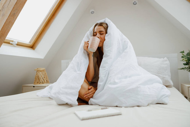 Una mujer está sentada en una cama envuelta en una manta blanca, sonriendo y bebiendo café - Foto, imagen