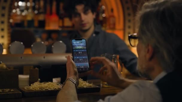 Mature sports fans check game score and bookmaker ratings in app using mobile phone, talk with bartender. Male friends bet on football match online sitting at bar counter in pub. Gambling concept. - Footage, Video