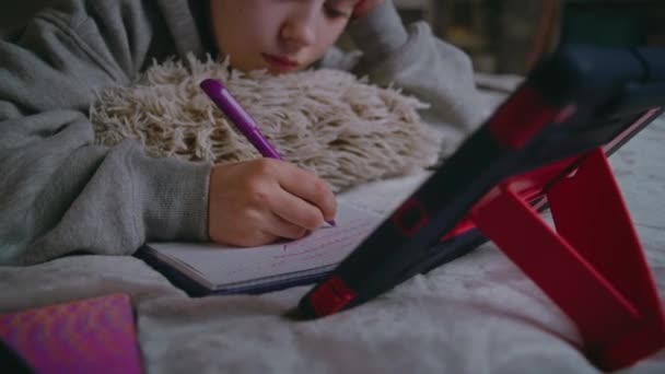 Primer plano de la joven chica caucásica escribiendo en el cuaderno, haciendo la tarea de la escuela en línea en el acogedor dormitorio usando la computadora de tableta. Adolescente pasa el tiempo y estudia acostado en la cama en casa. Concepto de estilo de vida. - Metraje, vídeo