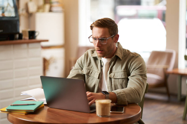 Portrét muže s vážnou tváří, sedí v kavárně, nosí brýle, pracuje na notebooku, vypadá soustředěně, dělá projekt na volné noze. - Fotografie, Obrázek