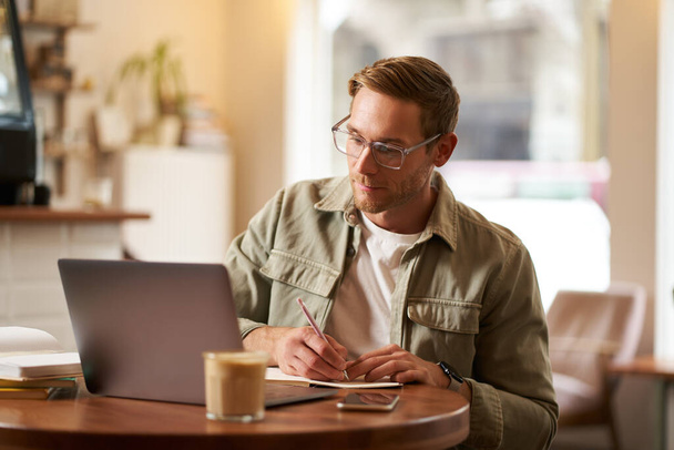 Image of focused young man in glasses, sitting in cafe, making notes, studying, attending online course, learning on remote from quite coffee shop. - Foto, imagen