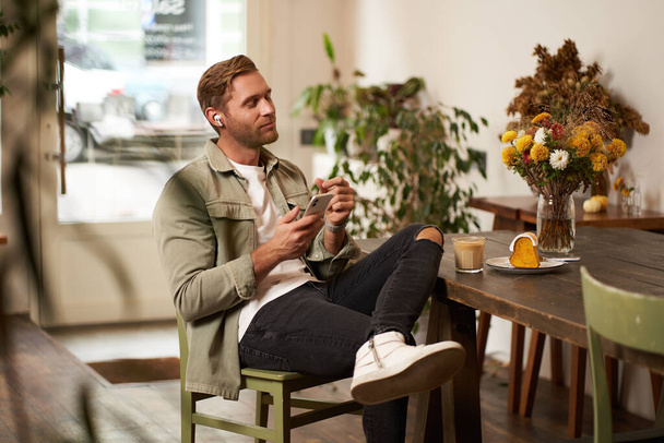 Portrait of young handsome man in casual clothes, sits in cafe and enjoys favourite song, listens to music in wireless headphones, vibing to the relaxing tunes in earphones, holding smartphone. - Foto, immagini