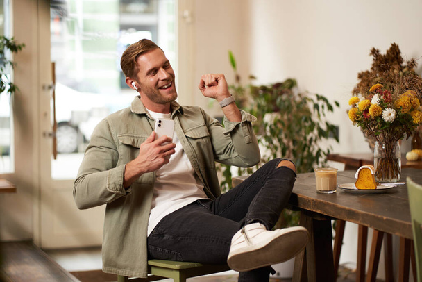 Портрет красивого счастливого человека в кафе, слушает музыку в беспроводных наушниках, держит смартфон, подключается к общественному Wi-Fi и наслаждается любимой песней, расслабляясь в кафе. - Фото, изображение