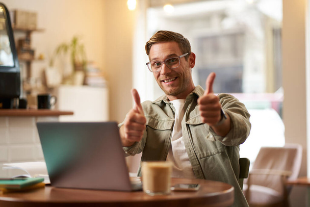 Радостный улыбчивый парень в очках, мужчина показывает большие пальцы вверх, сидит в кафе с ноутбуком, веб-сайтом, сервисом электронного обучения или коворкингом. - Фото, изображение