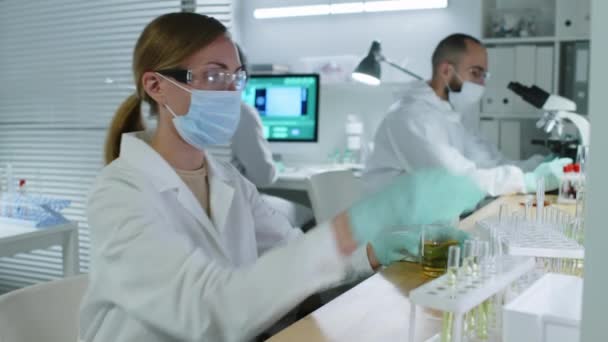 Aufstehen einer jungen kaukasischen Chemikerin mit Schutzbrille, Mundschutz und weißem Laborkittel, die während des Experiments im Labor unkenntliche Flüssigkeit aus einem Glaskolben in ein Becherglas gießt - Filmmaterial, Video