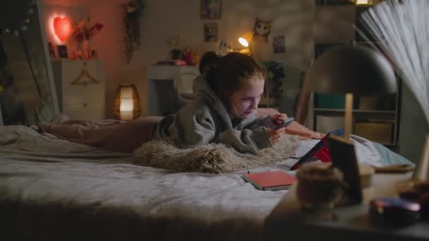 Caucasian teenager uses her mobile phone to surf Internet and chat with friends in social network. Young girl lies on big cozy bed and rests in evening. Teen spends time in comfort bedroom at home. - Footage, Video