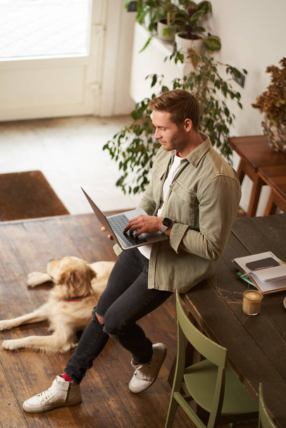 Κάθετη λήψη του όμορφου επιχειρηματία, νεαρός άνδρας με φορητό υπολογιστή ακουμπά στο τραπέζι, κάθεται σε χώρο συνεργασίας με το σκύλο του. Νεαρός πωλητής ελέγχει τα μηνύματά του, χαμογελά, εργάζεται σε ένα γραφείο με ένα κατοικίδιο ζώο. - Φωτογραφία, εικόνα