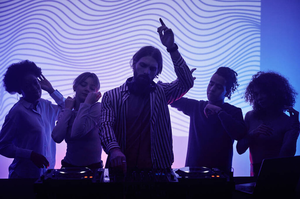 Съемка группы молодых людей, танцующих с диджеем в неоновом свете на вечеринке ретро-диско - Фото, изображение