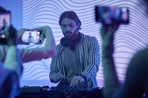 Portret długowłosego DJ-a przez stację muzyczną w neonowych światłach zabawiający tłum na imprezie disco - Zdjęcie, obraz
