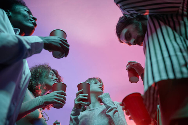 Low angle view at group of young people at house party holding red cups with alcohol and dancing in pink neon light - Photo, Image
