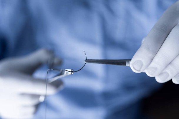 Un professionnel de la santé en blouse bleue manipulant des sutures. Opérateur tenant une aiguille demi-cercle avec pinces et porte-aiguille pendant le fonctionnement. - Photo, image