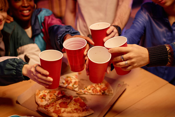 Nahaufnahme von Freunden, die rote Tassen in der Hand halten und bei einer Hausparty mit Pizza auf den Tisch anstoßen, Kopierraum - Foto, Bild