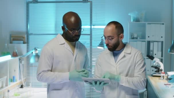 Colpo medio di due scienziati di sesso maschile etnicamente diversi che indossano camici e guanti monouso guardando tablet digitale e avendo conversazione mentre co-lavora sulla ricerca in laboratorio - Filmati, video