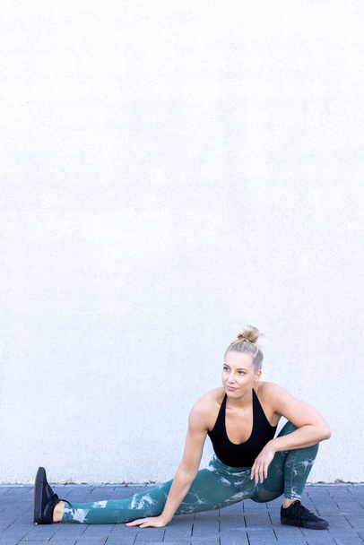 Siyah ve mavi spor elbiseli güzel sarışın bir fitness kadını, kaslarını ve kalçalarını hedeflemek için yere yakın bir ısınma egzersizi yapıyor ve sabah egzersizlerini geliştiriyor. - Fotoğraf, Görsel