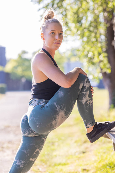 Atletik giyinen bir kadın, etkin bir yaşam tarzını vurgulamak için güneşli bir parkta bacağını esnetiyor. Sunlit Park 'ta esneme bacağı olan kadın. Yüksek kalite fotoğraf - Fotoğraf, Görsel