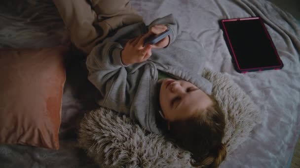 Jong meisje met koptelefoon ligt thuis op bed, luistert naar muziek en surft op internet met een mobiele telefoon. Blanke tiener brengt overdag vrije tijd door in de comfortabele slaapkamer. Levensstijl concept. - Video