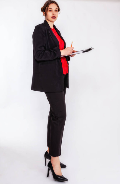 Een jonge, blonde vrouw met een broodje draagt een zwarte broek en een rode blouse. Ze houdt een schrijfbord en een potlood in haar hand en kijkt aandachtig in de camera. De achtergrond is grijs. - Foto, afbeelding