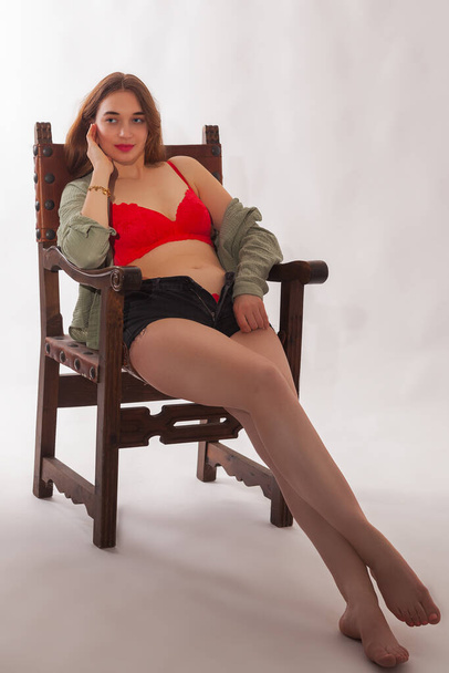 Een jonge, blonde vrouw met lang haar zit op een Spaanse stoel in een studio. Ze draagt een rode lingerieset bestaande uit hot pants en een top met lange mouwen en een off-the-shoulder halslijn. Ze lijkt zelfverzekerd en sensueel.. - Foto, afbeelding