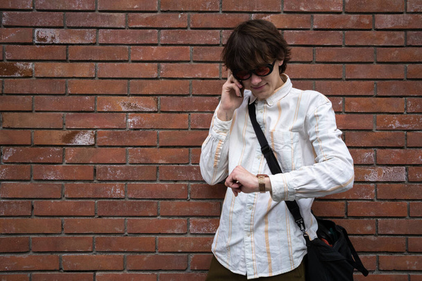 Joven hombre con camisa rayada chequeando reloj mientras habla por teléfono contra la pared de ladrillo. - Foto, Imagen