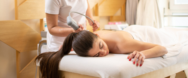 Extracorporeal shockwave therapy in beauty salon. Physical therapy for neck and back muscles,spine with shock waves. Young woman wearing protective mask during treatment in salon. - Photo, Image