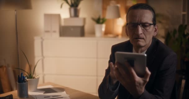 Grave senior manager di sesso maschile che legge sul tablet digitale mentre lavora fino a tarda notte in ufficio - Filmati, video