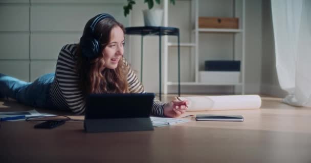 Mujer joven sonriente escuchando música mientras dibuja en el portapapeles tumbado en la sala de estar en casa - Metraje, vídeo